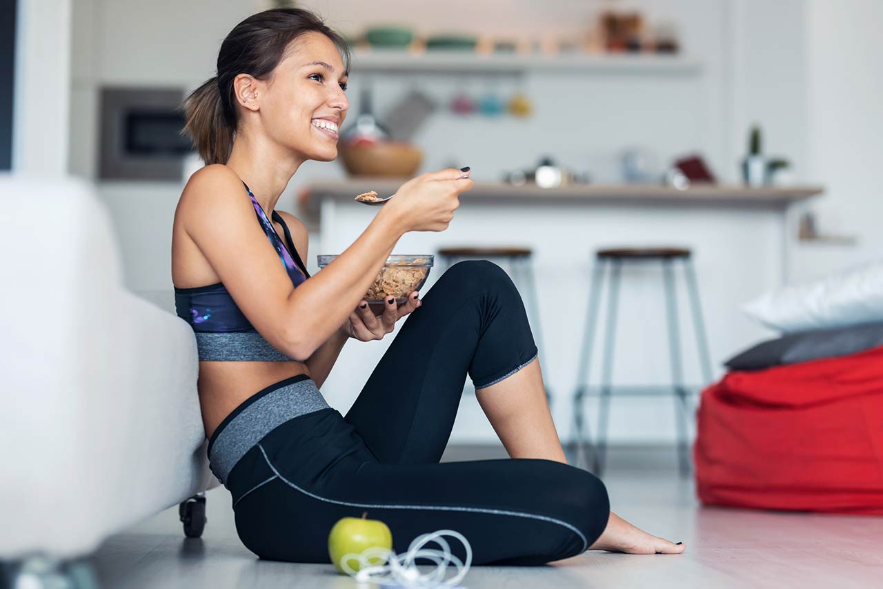 Sporty young woman eating a bowl of muesli while listening to music sitting on the floor at home