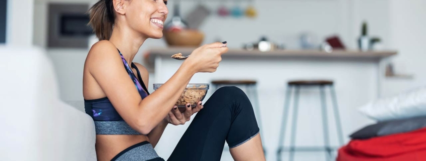 Sporty young woman eating a bowl of muesli while listening to music sitting on the floor at home