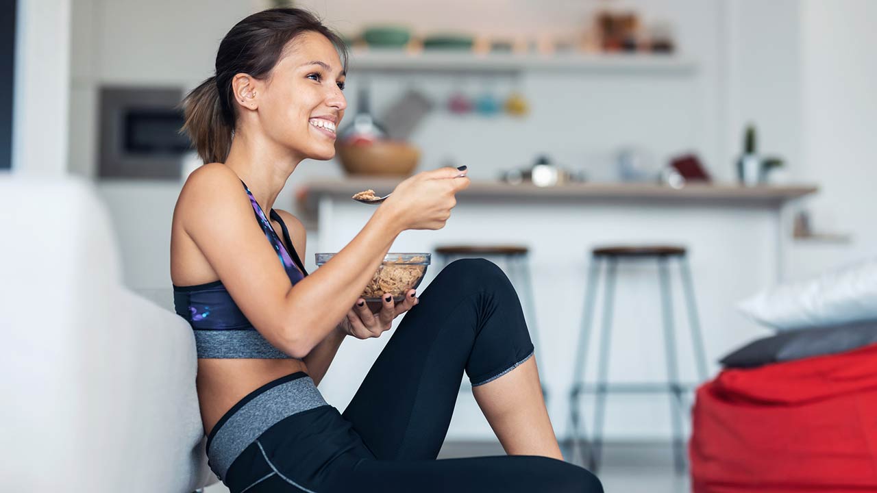 Sporty young woman eating a bowl of muesli while listening to music sitting on the floor at home