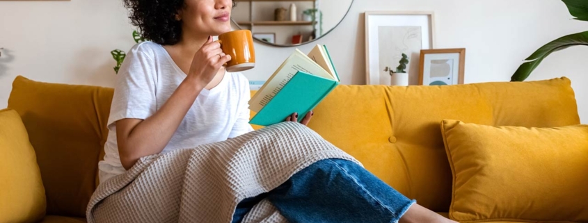 Relaxed African american woman reading a book at home, drinking coffee sitting on the couch