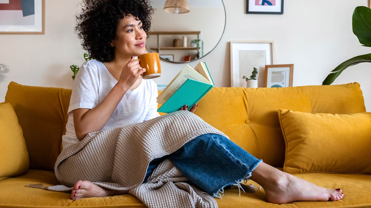 Relaxed African american woman reading a book at home, drinking coffee sitting on the couch