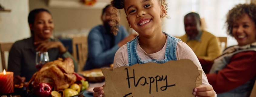 Happy African American little girl holding Happy Thanksgiving placard while celebrating with her family at home and looking at camera