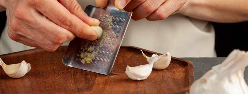 Hands of a caucasian woman grating garlic cloves using shovel shaped metal mini grater on a wooden plate