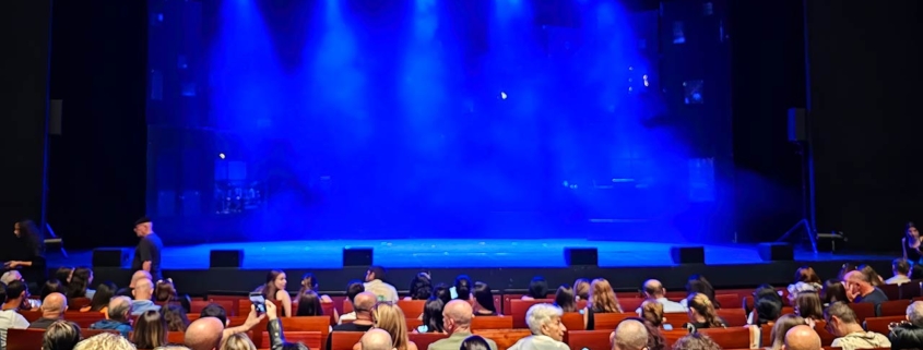An audience in theatre chairs with a stage and blue lighting in the background