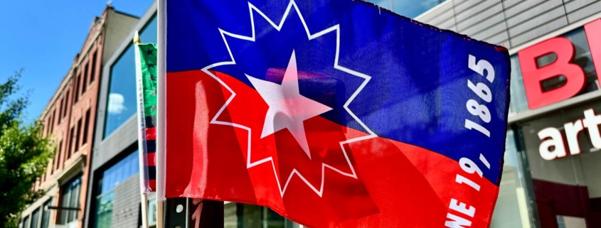 Small blue and red Juneteenth flag with a star and the date June 19, 1865 waving in the wind at a street vendor's stall in front of shops