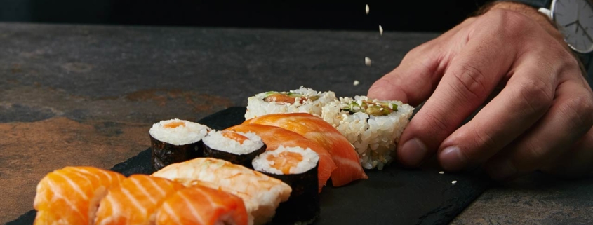 partial view of chef pouring sesame seeds on arranged sushi set on slate plate on dark tabletop