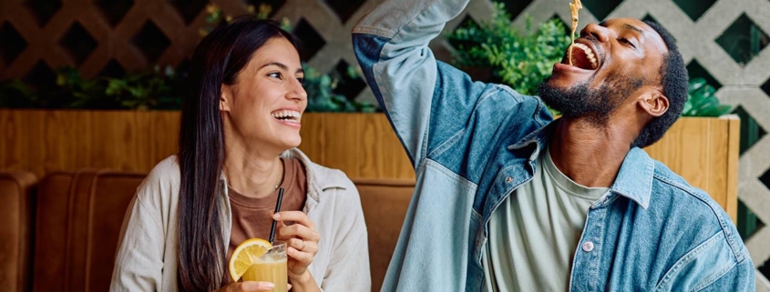 Cheerful multiracial couple laughing together during a restaurant dinner date, man playfully eating pasta, woman drinking a beverage