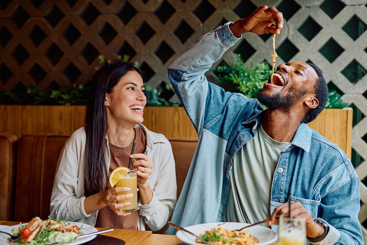 Cheerful multiracial couple laughing together during a restaurant dinner date, man playfully eating pasta, woman drinking a beverage