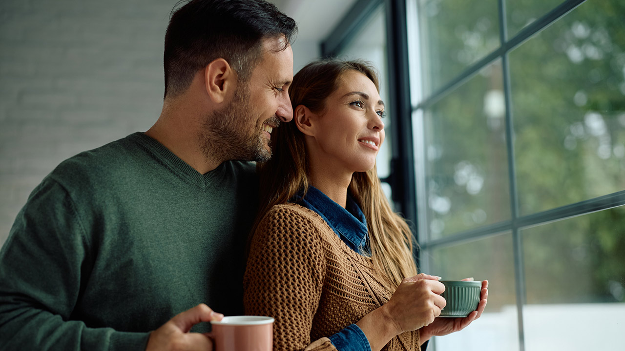 Smiling woman day dreaming while drinking coffee with her husband by the window at home