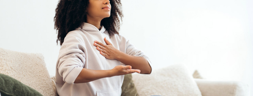 Serene young woman practicing breathing exercises while sitting in lotus position on comfortable sofa at home